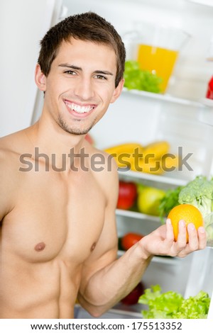 Man hands orange standing near the opened fridge. Concept of healthy and dieting food