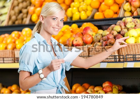Girl reads shopping list near the stack of fruits lying in the braided baskets in the store
