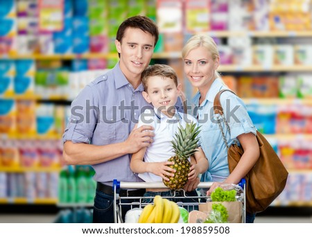 Half-length portrait of family in the shop. Son keeps pineapple over the shopping trolley full of food