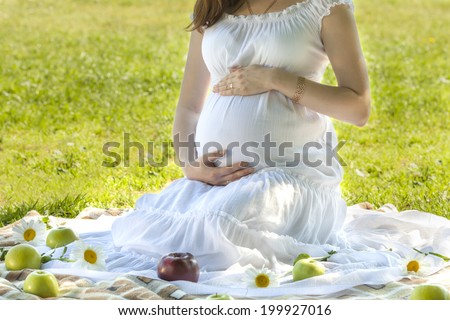 happy future mother sits on a lawn and hugs belly. In white dress