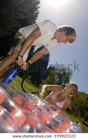 Young man inflating air bed, teenage girl holding hose, tilt view