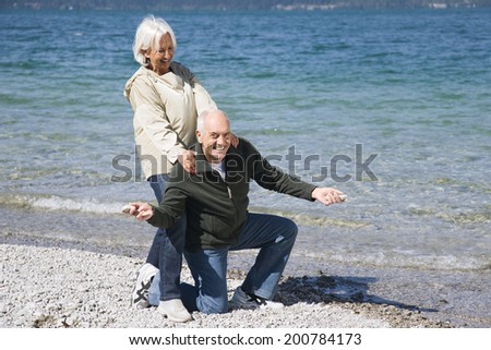 Germany, Bavaria, Walchensee, senior couple fooling about on lake shore man kneeling woman standing behind his back