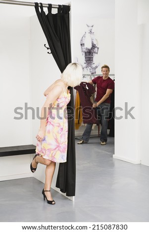 Young woman in changing room, man in background choosing dress