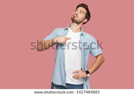 This is me. Portrait of proud haughty handsome bearded young man in blue casual style shirt standing, looking away and pointing himself. indoor studio shot, isolated on pink background.