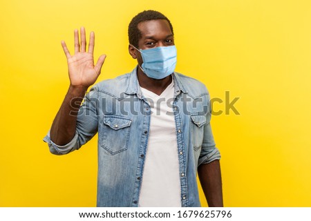 Hello! Portrait of positive handsome man with medical mask with rolled up sleeves smiling friendly and waving hand saying hi, welcoming gesture. indoor studio shot isolated on yellow background
