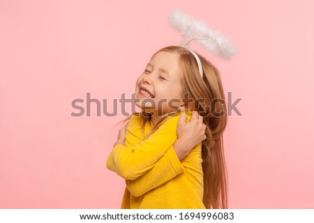 I love myself. Beautiful charming little girl with halo over head embracing herself and smiling from happiness, self-love concept, positive self-esteem. indoor studio shot isolated on pink background