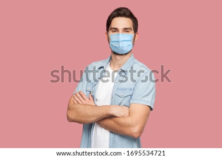 Portrait of handsome young man with surgical medical mask in blue casual style shirt standing, raised arms and looking at camera with smile. indoor studio shot, isolated on pink background.