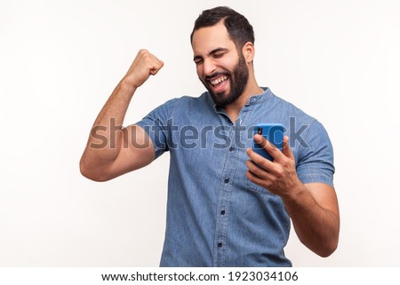 Happy satisfied man with beard in blue shirt holding smartphone and smiling making yes gesture, celebrating online lottery or giveaway victory. Indoor studio shot isolated on white background