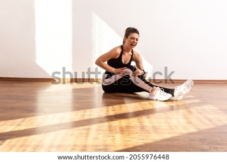 Sportswoman shouting from knee pain, touching leg, suffering kneecap injury during workout, wearing black sports top and tights. Full length studio shot illuminated by sunlight from window. Сток-фото © 