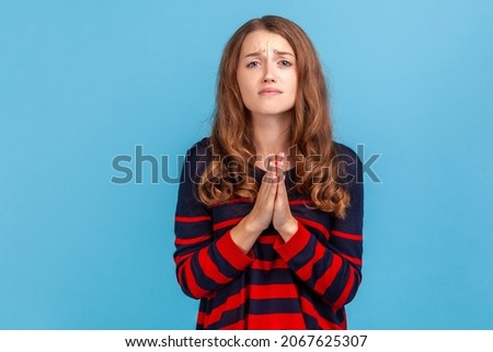Woman wearing striped casual style sweater holding hands in prayer, looking with imploring pleading expression, begging help, asking forgiveness. Indoor studio shot isolated on blue background. Imagine de stoc © 
