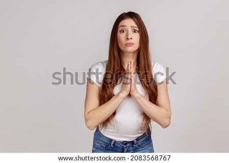 Please, I need. Young adult woman holding hands in prayer and asking for help with pleading imploring expression, full of hope, wearing white T-shirt. Indoor studio shot isolated on gray background. Imagine de stoc © 