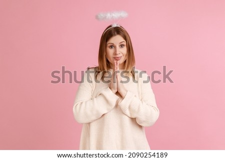 Portrait of blond woman with saint nimbus holding hands in prayer and looking with obedient imploring expression, asking help, wearing white sweater. Indoor studio shot isolated on pink background. Imagine de stoc © 
