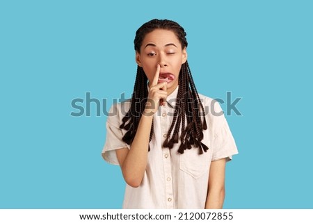 Portrait of funny impolite woman with black dreadlocks making crazy face with tongue out and picking her nose, bad manners, wearing white shirt. Indoor studio shot isolated on blue background. Foto stock © 
