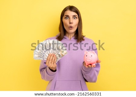 Portrait of shocked surprised woman with dark hair holding big sum of money and piggy bank, profitable investment, wearing purple hoodie. Indoor studio shot isolated on yellow background. Stok fotoğraf © 
