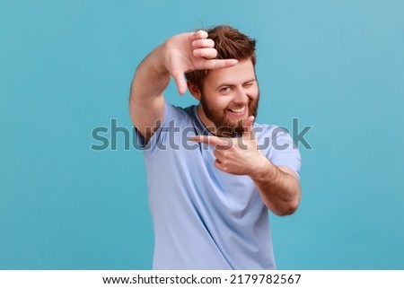 Portrait of curious bearded man gesturing picture frame with hands, looking through fingers and focusing on interesting moment, taking photo. Indoor studio shot isolated on blue background. Foto d'archivio © 