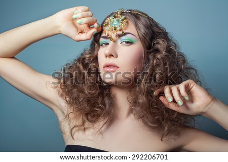 Beautiful fashion model with elegant gold middle eastern headpiece on head looking at camera with serious look and curly hair. on blue background.\
RAW - retouched with special care and attention.