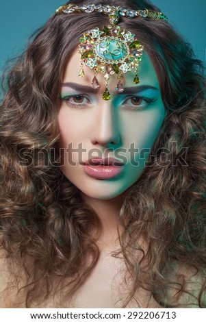 Beautiful fashion model with elegant gold middle eastern headpiece on head looking at camera with serious look and curly hair. on blue background.\
RAW - retouched with special care and attention.