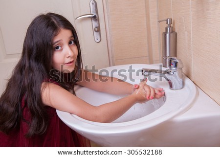 one beautiful 7-8 years middle eastern arab girl with red dress is washing her hands in the bathroom. looking at camera with smile. Developed from RAW. retouched with special care and attention.
