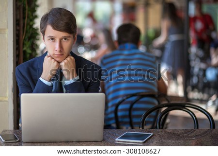 One relaxed young handsome professional businessman working with his laptop, phone and tablet in a noisy cafe. unhappy looking at camera.