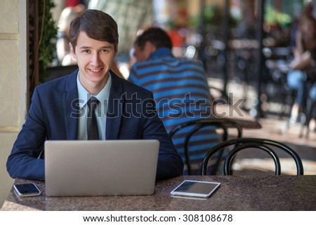 One relaxed young handsome professional businessman working with his laptop, phone and tablet in a noisy cafe. looking at camera and smiling.