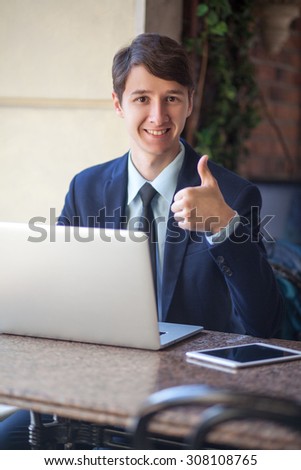 One relaxed young handsome professional businessman working with his laptop, phone and tablet in a noisy cafe. \
happy and looking at camera with smile and thumb up.