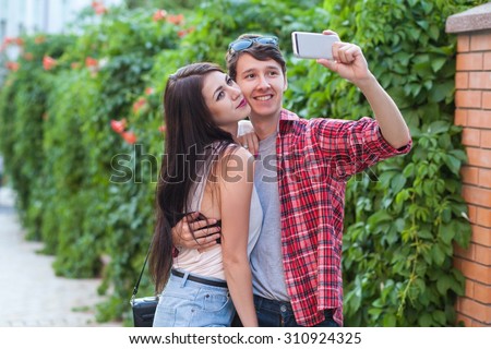 happy young tourists couple taking a selfie with smartphone on the stick in city. The man is holding the stick and shooting looking at phone with happiness. technology and friendship concept.