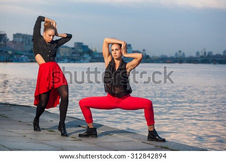 Two young beautiful twin sisters are dancing waacking dance in the city background near river. showing the different style and pose of modern dance with black and red dress near water on summer time.