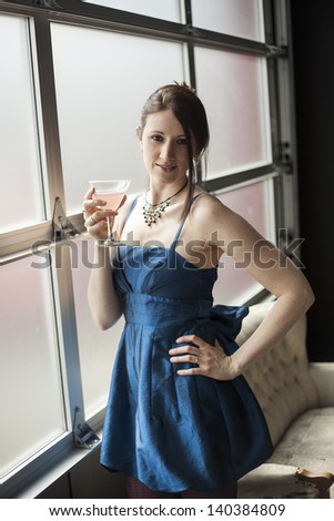 Beautiful young woman with brown hair and eyes in blue satin cocktail dress. She is drinking a pink martini.