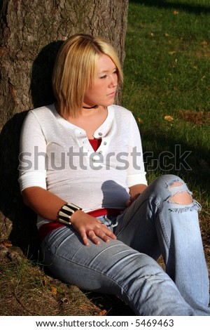 Serious young woman sitting on the ground near a tree, with her face turned to her left toward the sunlight.