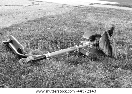 Black and white portrait of a scooter with an attached baseball glove and hat, and lunch box, laying on a lawn.