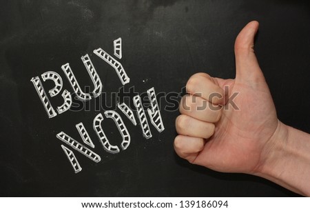 The words Buy Now in stencil lettering on a blackboard with a hand giving the Thumbs Up sign