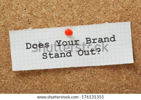 The phrase Does Your Brand Stand Out typed on a piece of graph paper and pinned to a cork notice board.