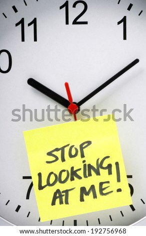 Yellow sticky note with the message Stop Looking At Me written on it and stuck to an office wall clock, as a concept for clock watching at work or school.