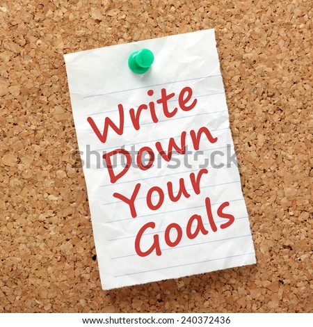 The phrase Write Down Your Goals written on a piece of paper and pinned to a cork notice board as a reminder
