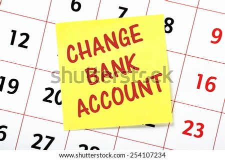 A reminder to Change Bank Account written in red ink on a yellow sticky note and pasted to a wall calendar
