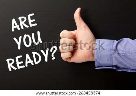Male hand wearing a business shirt giving the thumbs up gesture to the phrase Are You Ready in white text on a blackboard
