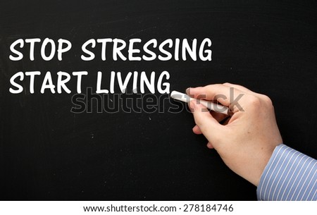 Male hand wearing a business shirt writing the phrase Stop Stressing Start Living in  white text on a blackboard
