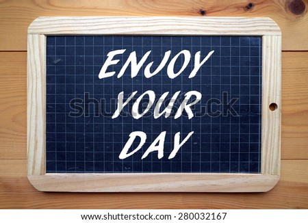 The phrase Enjoy Your Day in white text on a slate blackboard placed on a wooden surface