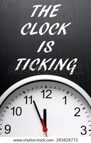 The phrase The Clock is Ticking in white text on a blackboard above a modern wall clock, with the hands at midnight or twelve noon