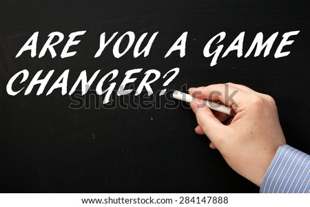 Male hand wearing a business shirt writing the question Are You A Game Changer in white text on a blackboard