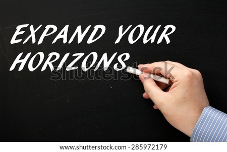Male hand wearing a business shirt writing the phrase Expand Your Horizons in white text on a blackboard