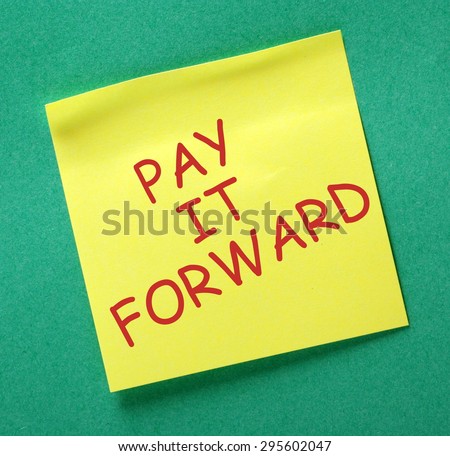 The phrase Pay It Forward in red text on a yellow sticky note posted on a green notice board