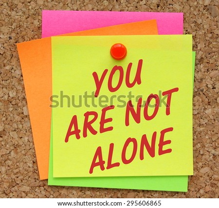 The phrase You Are Not Alone in red text on a yellow sticky note pinned to a cork notice board as a reminder