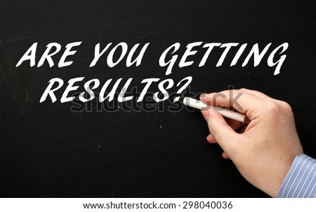 Male and wearing a business shirt writing the question Are You Getting Results? in white text on a blackboard