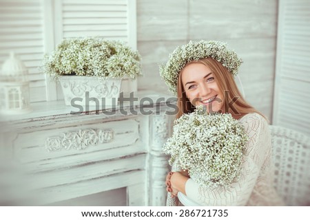 A happy girl is keeping flowers and grinning all over her face. She is sitting in the armchair near the fireplace. She is having a floral wreath on. The atmosphere of happiness is everywhere.