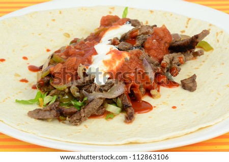 Flour tortilla on plate being filled with beef fajita; spicy pico de gallo; salsa; sour cream; onions; peppers; cheese and other ingredients of large burrito Mexican Dinner.