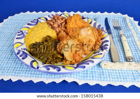 Soul food supper of fried chicken with collard greens; creamed corn; black eyed peas and candied yams on blue gingham background.