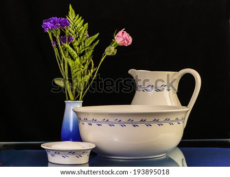 Antique Wash Basin with matching water pitcher and soap dish on glass-top wash stand with  flower arrangement in blue vase on black background with reflections and copy space.
