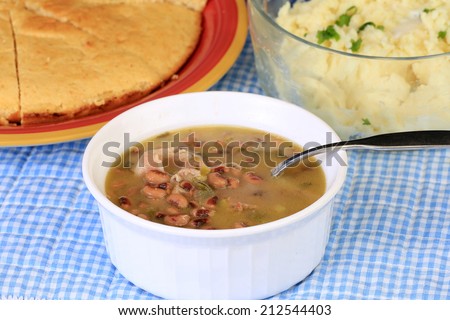 Cornbread and mashed potatoes fronted by bowl of black-eyed peas seasoned with fat back and bacon on blue gingham Southern Style setting.