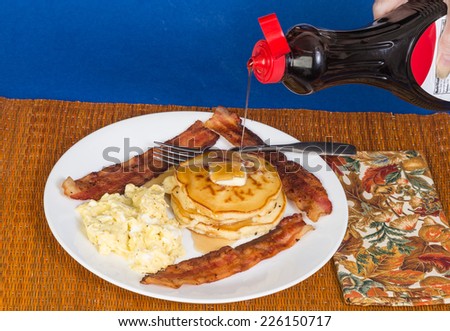 Scrambled eggs, fried bacon and stack of buttered pancakes with syrup being poured from plastic bottle.